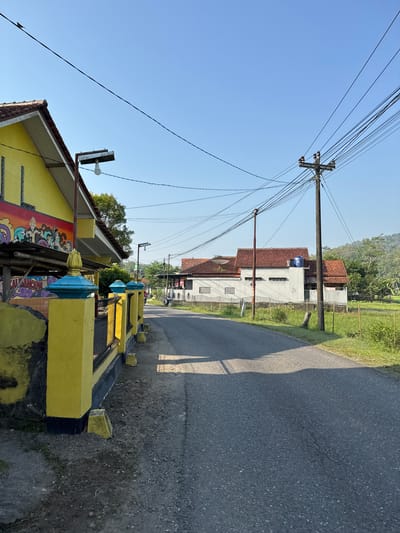 The image depicts a residential street scene in the village of Seliling, Indonesia, on a clear, sunny day. In the foreground, a bright yellow fence with blue decorative tops lines the left side of a paved road. Behind the fence, a yellow building with a red tile roof is visible, featuring a mural with figures and text that appears to be in Indonesian. The road itself is asphalt and curves slightly into the distance.

On the right side of the road, there is a grassy area with some vegetation and a rice paddy. Further down the road, several houses with red tile roofs and white walls are situated. Electrical poles with numerous wires crisscross the sky, a common feature in Indonesian infrastructure. One of the houses has a blue water tank on its roof. A small motorcycle is parked on the side of the road near one of the houses. The overall impression is of a quiet, rural or semi-urban setting with typical Indonesian architecture and infrastructure.