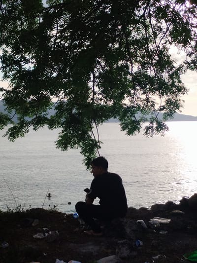A man is crouched down on the ground by the shore of a body of water, partially obscured by the dark silhouette of a tree hanging overhead. The man is dark-haired and wearing dark clothing, holding a phone in his hand. In the water are two dark shapes, possibly people swimming or objects. The ground near the man is littered with plastic waste. The scene is at Lamreh, Indonesia, judging by the location.