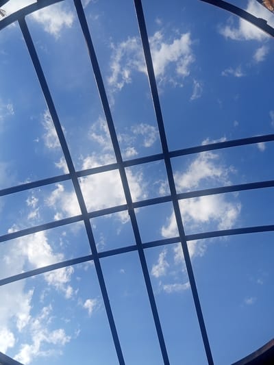 The image shows a view of the blue sky with scattered white clouds, framed by dark metal beams. The beams form a grid pattern across the sky, with some curved elements suggesting a roof or canopy structure. The perspective is looking upwards from below the structure. The scene appears to be outdoors on a clear day. There are no people or specific objects identifiable in the sky, only the natural elements and the man-made structure. There is no contextual information available to place this image specifically in Jardy-Su, Kyrgyzstan.