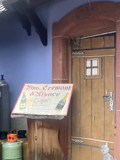 The scene depicts a portion of a building exterior in Kintzheim, France. The left side features a smooth, periwinkle-blue wall with a dark, carved wooden bracket extending from the upper left. To the right, a reddish-brown architrave frames a brown, paneled wooden door. The door is adorned with painted details, including a small, four-pane white window in its upper half, a stylized silver door handle and lock plate, and a silver horizontal decorative bar below the window. Green painted foliage is visible near the top left of the door, and a gray, cartoonish cat figure with white eyes is painted at the door's lower right. In the foreground, positioned centrally, is a weathered rectangular sign with a red border, reading "Vins & Crémant d'Alsace" in ornate script. The sign illustrates two wine bottles, one with a blue label on the left and one with a yellow label on the right. Additional text on the sign references "Hotel & Jean-Michel KOENIG" and "Crée direct du Producteur au Consommateur Cartons de 3.6.12 Bouteilles". This sign rests on a dark, curved support structure. To the immediate left of the sign, partially visible, are several gas cylinders: a tall, light gray cylinder with a black valve at the top and multiple smaller, green cylinders with orange caps in the lower foreground. No people are present in the image.