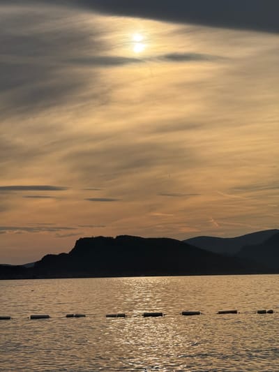 The image depicts a serene seascape at sunset, featuring a body of water with a faint line of buoys in the foreground. A silhouetted mountain range forms the horizon, with a prominent rocky outcrop on the left. The sky above is filled with streaky clouds, through which the setting sun's golden light filters, creating a warm and atmospheric glow. The location is identified as Košljun, Montenegro.