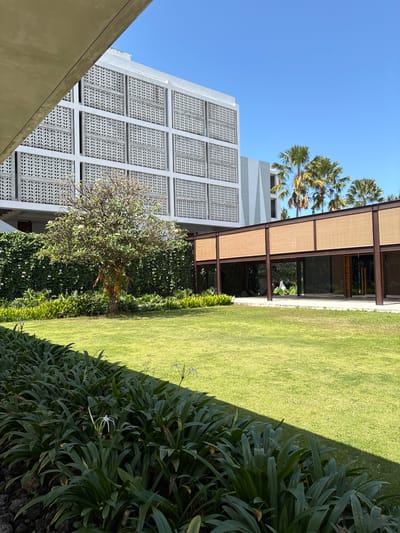 This image depicts the exterior of a modern building with a lush green lawn in the foreground. The building features a distinctive facade with patterned concrete panels and a covered walkway with woven rattan accents. Palm trees are visible in the background. The scene is set under a clear blue sky. The perspective suggests this is likely a hotel or resort in Banyuwangi, Indonesia, given the architectural style and tropical vegetation.