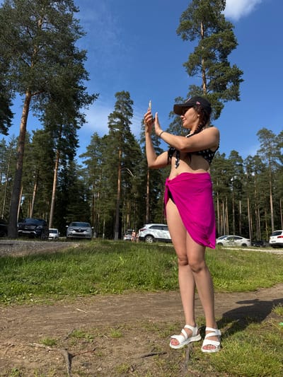 In an outdoor scene, a woman stands in a sun-drenched forest clearing, holding a mobile phone to take a photograph. She wears a black and white polka-dot bikini top, black bikini bottoms, and a vibrant fuchsia sarong tied around her waist. White strap sandals adorn her feet. Her hair is in a braid, and she wears a black baseball cap. The background reveals a line of parked cars, including black, silver, and white vehicles, suggesting a public access area or a gathering spot. Tall pine trees with dense green foliage dominate the landscape, their trunks a reddish-brown. The ground in the foreground is a mix of dry dirt and patches of green grass. The sky above is a clear, bright blue with wispy white clouds. The scene is set on a sunny day, with dappled shadows indicating the sun's position overhead. The location is identified as Korkino, Russia, likely a recreational area near a forest.