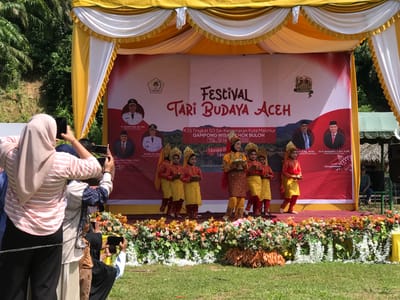 This image captures an outdoor cultural festival event. The main focus is a stage decorated with yellow and white fabric draping and adorned with a vibrant floral arrangement at its base. A large red banner serves as the backdrop, featuring event details written in Indonesian, including "FESTIVAL TARI BUDAYA ACEH" which translates to "Aceh Cultural Dance Festival." The banner also displays official seals and portraits of individuals in formal attire, likely dignitaries.
On the stage, several young performers dressed in traditional Acehnese attire are present. They wear colorful costumes consisting of embroidered tops and pants or skirts, often accompanied by decorative headwear. Some of the performers are holding small bowls.
In the foreground, a crowd of spectators is visible, some of whom are holding up smartphones to capture the event. The ground in front of the stage is covered in green grass, and lush green foliage and trees are seen in the background, suggesting a natural or semi-rural setting. The overall atmosphere appears festive and celebratory. The specific location is identified as Pantonrayeuk Satu, Indonesia, indicated by text on the banner.