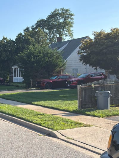 A medium shot captures a suburban residential street scene in Levittown, United States of America. The foreground shows a paved road and a sidewalk bordering a well-maintained lawn. Beyond the lawn, a light-colored wooden fence runs parallel to the road, partially obscuring a gray trash bin labeled "BRUTE".
Two red vehicles are parked in the driveway of a single-family home. The car closer to the foreground is a Dodge Challenger, while the car further back appears to be a Mazda CX-5. The house itself is a light-colored, two-story dwelling with a dark-pitched roof. A white gazebo with decorative pillars stands in the front yard, partially obscured by dense green shrubbery. Lush green trees of varying sizes surround the property, and the sky above is a clear, bright blue. The image appears to be taken on a sunny day. There are no people visible in the frame.
