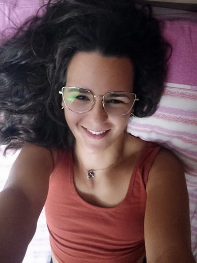 This image depicts a single female individual in an indoor setting, likely a private residence in San Marzano sul Sarno, Italy. The subject is lying on her back on a bed, viewed from a high-angle perspective, consistent with a selfie. Her long, dark, wavy hair is fanned out around her head, resting on the bedding. She wears gold-rimmed, cat-eye style eyeglasses, with reflections visible in the lenses. Her facial expression is a broad, open-mouthed smile, revealing both upper and lower teeth. She is wearing a rust-orange colored tank top. A thin, gold-toned chain necklace with a small, dark, possibly heart-shaped pendant is visible on her upper chest. The bedding consists of a plain pink or light purple section near her head, transitioning to a white sheet with thin, horizontal pink and purple stripes beneath her body and torso. Her right arm (viewer's left) is extended slightly towards the bottom left of the frame. No other subjects or distinct objects are visible in the immediate background.