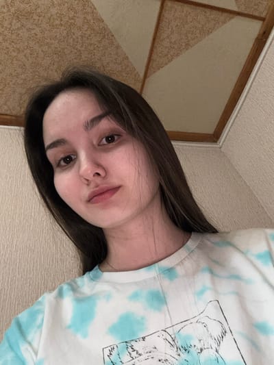 An indoor, close-up image taken in Bayronovka, Russia, features a young female subject looking directly into the camera. The subject has fair skin, dark brown hair parted centrally, and a neutral expression. She wears a white t-shirt with a light blue tie-dye pattern and a partially visible dark graphic print on the lower right depicting what appears to be a figure. The background includes a light-colored, possibly textured wall to the left and a ceiling featuring a geometric pattern of light-toned panels with a speckled texture, framed by dark wooden or faux wood strips. The ceiling and wall meet at a corner defined by a wooden trim. The lighting is consistent with an interior setting.