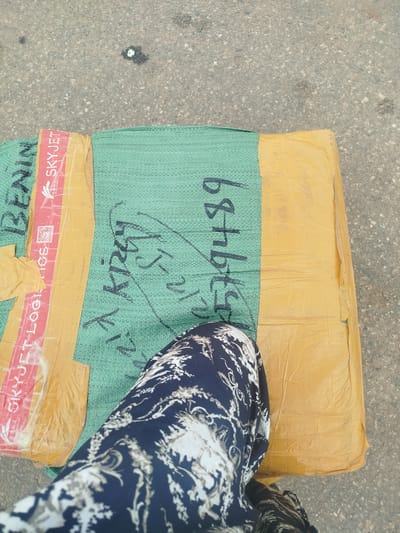 The image shows a package resting on a paved surface, possibly a road or sidewalk. A person's legs, clad in dark blue patterned trousers, are visible in the foreground, partially obscuring the package. The package is wrapped in a yellow paper material and covered with a green woven fabric. Red and white tape bearing the text "SKYJET LOGISTICS" is visible on the left side of the package. Black handwriting is present on the green fabric, including what appears to be a date or tracking number: "15 2019" and other illegible scribbles. The paved surface suggests an outdoor setting. The overall lighting implies it is daytime. The location is identified as Uselu, Nigeria.