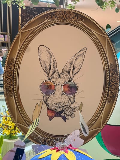 An oval-framed artwork depicting a rabbit is centrally positioned in an indoor commercial setting within Vösendorf, Austria. The frame is gold-colored with ornate, classical relief patterns along its border. The artwork itself features a black-and-white sketch of a rabbit's head and upper chest on a light beige background. The rabbit wears round, iridescent sunglasses, a small red bow tie, and holds a white multi-petaled flower in its mouth.
In the foreground, below the artwork, various decorative elements are visible. These include a prominent blue, possibly egg-shaped object with yellow felt petals and coiled silver wire accents, and several cardboard cutouts resembling abstract wings or feathers, attached to wires. To the left foreground, a small potted plant with vibrant yellow flowers is present.
The background shows an expansive interior space. Overhead, strands of artificial green foliage are suspended. Reflections on the artwork's surface indicate ambient light and architectural features of the environment, including a vertical element resembling an escalator on the right side. To the left, a dark, vertically oriented retail display or architectural feature is visible, along with a blurred, partially visible individual in the far left background. No explicit actions or interactions are depicted beyond the static display elements.