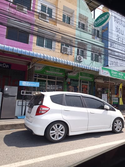 The image shows a white Honda Fit parked on a street in Hua Hin City Municipality, Thailand. In the background, there are several buildings with colorful facades. One building is painted with horizontal stripes of pink, beige, and green, while another is blue and has a green sign that translates to "Drugstore." There are numerous overhead power lines visible. A "Casper Electric" sign is displayed in front of a store that seems to sell appliances. The "PUNYA PLUS Pharmacy" is present, with green signs indicating the pharmacy's name in both English and Thai, along with contact information. The pharmacy appears to be open and selling pharmaceutical products. The overall setting appears to be a busy commercial street.