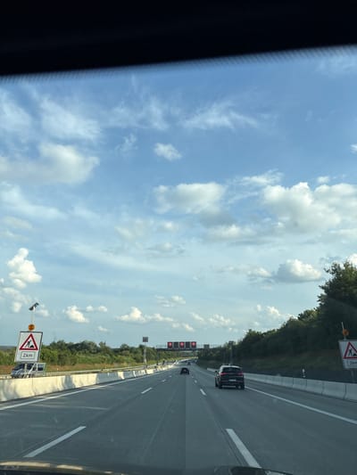 The image depicts a view from inside a vehicle traveling on a highway in Bindlach, Germany. The sky is partly cloudy with scattered white clouds against a blue backdrop. On the right side of the highway, trees and dense foliage form a natural barrier. Several traffic signs are visible, including a construction warning sign indicating "2 km" on the left and a similar sign on the right. Overhead gantries with speed limit signs (120 and 100) are present in the distance. A black SUV is traveling in the right lane, ahead of the viewer's vehicle. The highway has multiple lanes marked with white lines.