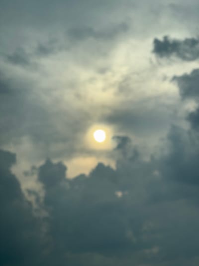 The image is a close-up, low-angle, blurred shot of the sky, taken from the city of Brashlen, Bulgaria. The sun is visible as a bright, out-of-focus orb in the upper center of the frame, partially obscured by dense, dark clouds. The clouds are textured and appear to be moving, creating a sense of depth and atmospheric movement. The overall lighting is dim, suggesting an overcast or stormy day. There are no people or discernible objects in the image, only the sky and clouds. The color palette is dominated by shades of grey and a pale yellow from the sun.