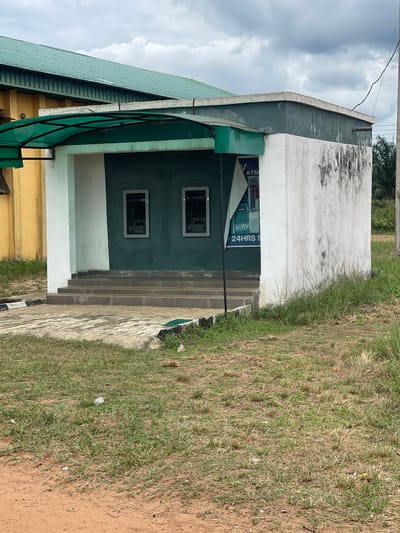 The image shows the exterior of a building in Abraka, Nigeria. The building appears to house an ATM, as evidenced by a sign reading "ATM" and "24HRS S" visible on the structure. The building's facade is a combination of green and white paint with a covered entrance featuring a green corrugated metal roof. There are two rectangular window openings on the front. A set of stairs lead up to the building's entrance. The surrounding environment consists of grass and dirt. The sky is overcast with cloudy conditions.