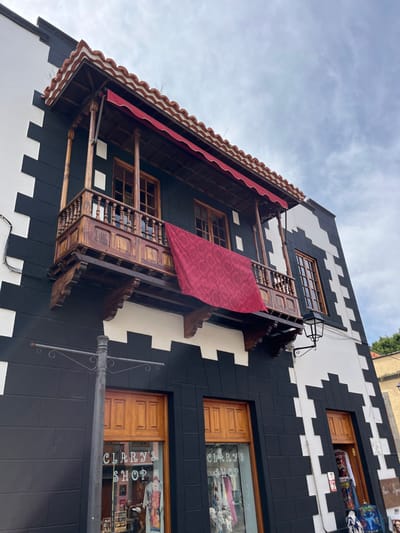 The image depicts a two-story building with a black and white painted exterior, located in Teror, Spain at coordinates 28.06, -15.547. The building has a prominent balcony extending out from the front of the second floor, which is made of wood and painted brown. There is a red awning over the balcony and another, larger red fabric draped over the balcony's railing, hanging down to the first floor. The balcony has a wooden railing and a metal streetlamp. The building has two storefront windows, one on each side of the balcony, both displaying the name "Clarry's Shop" in white letters on a black background. The windows are made of glass and have wood frames. The lower level of the building appears to be a commercial storefront. The scene is taken from a street-level perspective, looking up at the building. The sky is cloudy and the weather appears to be mild.