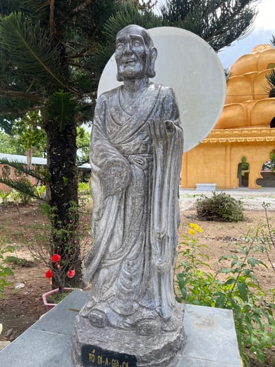 A weathered gray stone statue of an aged male figure stands prominently in an outdoor setting in Đăk Mar, Vietnam. The statue, identified by the inscription "TỔ DI-A-GIA-CA" on its base, depicts a robed individual with an aged face featuring prominent wrinkles, a beard, and a slightly open mouth. The figure's right hand is raised with an open palm, while the left hand is held closer to the body. Behind the statue's head, a large, flat, circular white disc is affixed. To the right of the statue, in the mid-ground, a multi-tiered golden-yellow Buddhist temple structure with decorative arched openings is visible. To the left and partially behind the statue, a tall coniferous tree with a dark trunk and green needles is present. The ground around the statue consists of exposed soil, sparse green vegetation, and small plants; a pot containing red-flowered plants is visible to the left, and taller green plants with yellow flowers are to the right. The scene is illuminated by natural daylight under a lightly clouded sky.