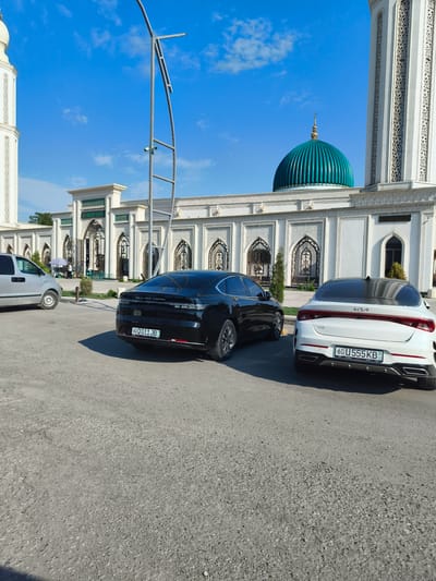 The image shows the exterior of the Azam-o-Rafiq Mosque in the city of Buz, Uzbekistan, on a clear, sunny day. In the foreground, two cars are parked on an asphalt lot. The car on the left is a black sedan with license plate "60 Q 011 JB". The car on the right is a white Kia sedan with license plate "60 U 555 KB". Behind the cars is the mosque's facade, featuring white walls with intricate decorative patterns and arched doorways. A large, teal-colored dome is visible atop the mosque, topped with a crescent moon. A tall, white minaret is also part of the mosque's architecture, located to the left of the dome. A modern-looking street lamp structure stands in the middle ground. In the background, a few trees and other structures are partially visible. The sky is bright blue with a few wispy clouds.