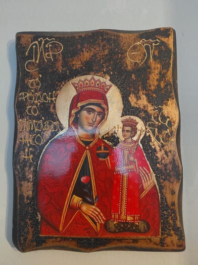 The image is a photograph of an icon on a wooden board. The icon depicts the Virgin Mary holding the Christ child. Both figures are wearing crowns, and Mary is holding a rose. The background is dark and textured, with gold lettering and symbols. The wooden board has a distressed, aged appearance. The icon is mounted on a wall, which is white and appears to be indoors. The photograph was taken in the Municipal Unit of Aegio, Greece.