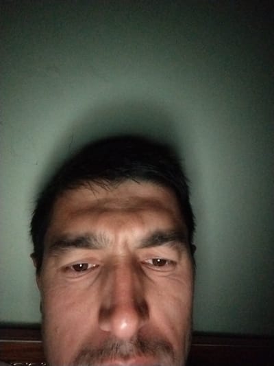 This is a close-up, low-angle selfie of a man's face, taken from the shoulders up, in what appears to be a dimly lit room with a green wall. The man has dark hair and brown eyes. He is looking directly at the camera with a neutral expression. His forehead shows some wrinkles. A faint shadow is cast on the wall behind him, suggesting a light source from the front and slightly above. The bottom edge of the image shows a dark horizontal line, possibly the top of a piece of furniture. There is no indication of Istaravshan, Tajikistan in the image itself.