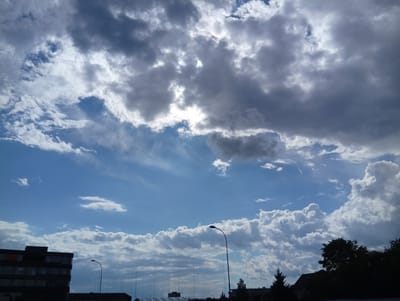 The image captures a view of the sky over Langenthal, Switzerland, with a mix of dark and light clouds against a bright blue sky. A silhouetted building, appearing to be an apartment complex or office building, is visible on the left side of the frame. A streetlamp with a curved arm stands in the middle ground, its light fixture facing upwards. In the distance, more trees and structures are faintly discernible, also in silhouette. The overall lighting suggests it is daytime, possibly late afternoon or early evening, given the contrast between the bright sky and the shadowed elements. The environment appears to be urban or suburban, with infrastructure like buildings and streetlights. There are no people or animals visible in the frame.