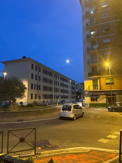 The image shows a street scene at dusk in Novi Ligure, Italy. A silver compact car is turning at a roundabout. To the left is a building with a light-colored facade and dark windows, and a tree in front of it. To the right is a taller building with balconies and windows, with a street lamp in front of it illuminating a storefront that sells fruits and vegetables. The sky is a deep blue, and a full moon is visible. There are other cars and people in the background. The scene suggests a typical urban environment in the evening.