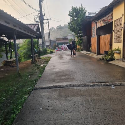 The image shows a street in Kecamatan Tamansari, Indonesia, at latitude -6.635, longitude 106.737. The street is paved with concrete and is wet, suggesting recent rain.  Two people are standing on the street, both wearing dark clothing.  One person is holding a black umbrella.  There is a child with them, also wearing dark clothing.  A group of people are visible further down the street, sitting on the sidewalk.  The street is flanked on both sides by buildings. The buildings on the left side are multi-story, with visible roofs, while the building on the right is a single-story, with a wooden gate and a brown fence. There are plants and shrubs growing in front of both buildings. The image appears to be taken from the street looking towards the buildings.