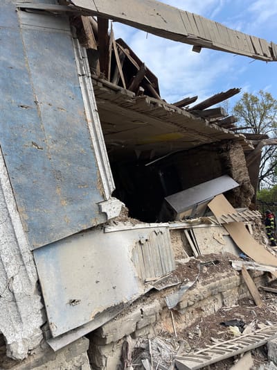 The image shows a collapsed building in Bilopillia, Ukraine. The building is made of brick and wood. The roof has collapsed, and debris is scattered throughout the scene. A large wooden beam is lying across the top of the building, with a piece of the roof still attached to it. There is a dark-colored cabinet, partially visible, inside the building. The scene is chaotic and appears to be the result of a destructive event, possibly an explosion or a military strike. A person in a red jacket, presumably a firefighter, can be seen in the bottom right corner of the image.