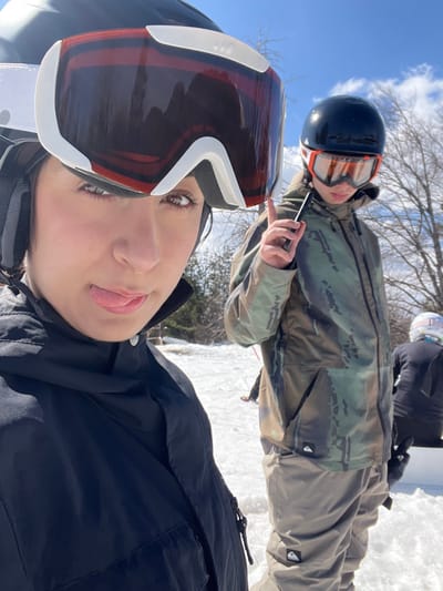 This image depicts an outdoor scene in the city of Bogoslov, Bulgaria, characterized by a snow-covered landscape under a clear blue sky with scattered white clouds. Three individuals equipped for winter sports are visible.

In the immediate foreground, a person with dark hair is partially shown, looking directly towards the camera with their tongue slightly extended. They are wearing a black winter jacket and a black ski helmet with white-framed goggles, featuring reddish-brown tinted lenses pushed up onto the helmet.

To the right of the foreground subject, in the midground, stands a second person, facing generally towards the right. This individual wears a camouflage-patterned winter jacket and khaki-colored snow pants, both items bearing a black square "Quiksilver" logo. They are equipped with a glossy black helmet and orange-framed, clear-tinted ski goggles. Their right hand holds a dark-colored smartphone vertically, with the index finger of the same hand raised.

In the background, positioned further to the right, a third individual is partially visible from behind. This person wears a dark-colored winter jacket and a white-framed helmet with pink-tinted goggles pushed up. They appear to be seated on a white structure.

The environment consists of snow-covered ground, and bare deciduous trees are visible in the distant background, consistent with a winter or early spring setting.
