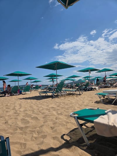 The image depicts a crowded beach scene under a bright blue sky with scattered clouds, likely taken in the city of Dyuni, Bulgaria. Numerous green beach umbrellas provide shade over rows of empty and occupied lounge chairs arranged on golden sand. Several people are visible, some seated on lounge chairs, others standing or walking, with a few in swimwear. The foreground features a close-up of a green lounge chair with a white towel draped over it, casting a distinct shadow on the sand. In the background, a gentle slope of hills covered in vegetation can be seen leading down towards the sea, which is not directly visible but implied by the beach setting. The overall atmosphere suggests a sunny day at a popular tourist destination.