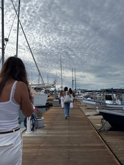 Here's a description of the scene:

The image shows a pier in La Paz, Mexico, filled with several sailboats. The perspective is oriented along the length of the pier. A group of people are walking along the pier. In the foreground, a woman with long hair is visible, wearing a white tank top and white pants with a brown belt. The sky is overcast with a layer of cloud cover. The sailboat "Cubana" is seen docked on the pier. A "Beneteau" sailboat is also visible. A gray flowerpot with pink flowers sits on the side of the pier. The pier is constructed of wooden planks.