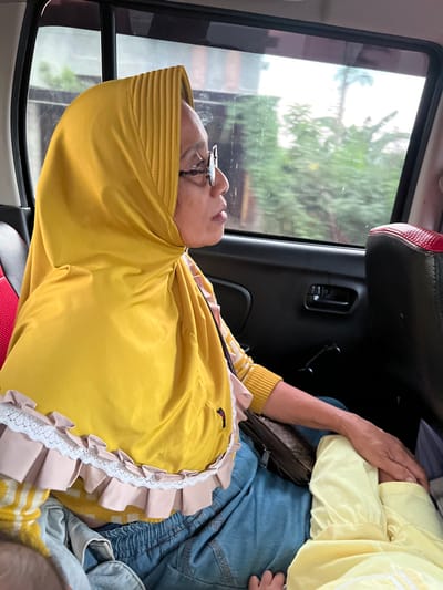 The image captures the interior of a vehicle, specifically the rear passenger compartment, in Terban, Indonesia. An adult female, appearing middle-aged or elderly, is seated in the left-hand rear passenger seat, viewed from her chest up. She wears a mustard yellow hijab featuring vertical pleats on the crown and decorative light peach ruffles with white lace trim along its lower edge. She has eyeglasses with thin frames and is looking out the window to her right. Her visible attire includes a yellow and white patterned long-sleeved top with ribbed cuffs, and blue pants with an elasticized waistband. A dark, possibly brown, pouch or bag rests on her lap. To her right, a second, smaller individual, likely a child, is partially visible; only their lower legs and a small hand are discernible, and they are dressed in light yellow clothing. The adult female's right hand rests on the child's leg. Through the vehicle's tinted rear window, blurred green foliage and indistinct background elements are visible, suggesting an outdoor environment. A black car door panel with a handle and a red and black upholstered headrest are also visible within the vehicle's interior.