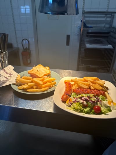 Two plates of food are presented on a metallic surface, likely a counter in a restaurant or catering establishment. The plate in the foreground contains a sandwich, cut diagonally and served with a generous portion of french fries. The sandwich appears to be toasted, with a filling visible. Below the plate, several crumpled receipts or order slips are partially visible. The plate in the background holds a salad with lettuce, red cabbage, cherry tomatoes, hard-boiled egg slices, and two sausages, accompanied by french fries. The lighting suggests it is daytime, possibly indoors, with some natural light filtering in, as evidenced by the reflections on the metallic surfaces. The overall scene is functional and practical, focused on the food presentation. The location context provided is Rivesaltes, France, but this is not directly inferable from the visual information alone. There is visible text on the receipts, but it is too blurry to be legible.