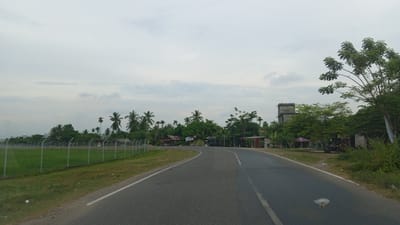 This is a photograph of a paved road in a rural area, likely near Keupok Nibong, Indonesia. The sky is overcast and filled with pale grey clouds. The road curves to the left, with a grassy shoulder on the left side of the image. A chain-link fence runs along the left side of the road, enclosing a green field. Beyond the field, there are numerous palm trees and other foliage, with several small buildings and structures visible amongst them. One building appears to be a multi-story concrete structure, possibly an industrial building or tower. The right side of the road is lined with vegetation and more trees, with some visible structures tucked behind them. The overall scene depicts a somewhat overcast day in a tropical or sub-tropical rural setting.