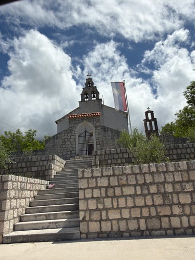 The image captures a serene outdoor scene, primarily featuring a traditional stone church or chapel nestled on a hillside under a bright, partly cloudy sky. The perspective is slightly upward, showcasing the architectural elements against the expanse of the sky.
The central subject is a stone-built church, characterized by its robust construction from irregular, light-colored stone blocks and a gabled roof covered with reddish tiles. An arched entrance, fitted with a dark, ornate wrought-iron gate, is visible on the side of the church. A prominent bell tower rises from the structure, featuring openings that house multiple bells, and is crowned with a cross. In the background, further up the hill, another, taller stone bell tower with a cross is partially visible, hinting at a larger complex or another religious edifice.
A flag flies from a pole situated near the main bell tower. It is a tricolor flag with horizontal stripes of red, blue, and white. Centered on the blue stripe is a golden emblem, which appears to be a stylized cross, possibly a Serbian cross (four C's/firesteels), indicative of historical and religious symbolism often found in the region.
To the right of the church, a robust stone wall, constructed from similarly irregular blocks, forms the base of a concrete staircase. Several concrete steps are visible, leading upwards along the right side of the image. Adjoining this stone wall on the far right is a smooth, light-colored concrete or stucco wall. Green trees and bushes are scattered throughout the scene, adding natural foliage around the stone structures.
The sky, occupying a significant portion of the image, is a vibrant blue, adorned with numerous fluffy white clouds, suggesting clear, bright daylight. No people or specific events are depicted, lending a tranquil and timeless quality to the scene. The architecture and flag strongly suggest a location in the Balkan region, specifically Bratešići, Montenegro, as provided by the context, reflecting the area's rich cultural and religious heritage.