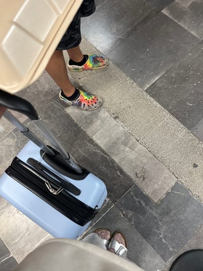The image depicts a first-person perspective view of two individuals at an airport or transport hub. One individual is wearing tie-dye Crocs with black socks, camouflage shorts, and is pulling a beige suitcase. The other person is wearing silver sandals, light pants and appears to be pulling a light blue suitcase. The floor is composed of dark and light gray marble tiling with a concrete pathway. The specific location in Puerto Escondido, Mexico, is not discernible from the provided image.