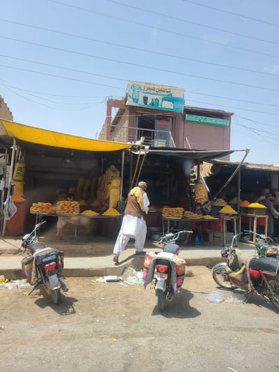 The image depicts an outdoor market scene in Saranan, Pakistan. A man in a brown and white outfit stands near a food stall, which is covered by a yellow awning. The stall displays various food items, possibly snacks, piled in baskets and bags, with one person visible inside the stall. Two motorcycles are parked on the dusty street in front of the stall, one with a colorful cover. The sky is clear and blue, suggesting a sunny day, likely in the morning or afternoon. A few signs are visible on the buildings behind the stall, one with Arabic script and another with an image of a woman. The overall atmosphere appears to be a typical day in a bustling local market.