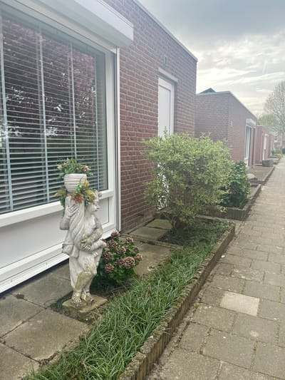 A photograph taken in Maastricht, Netherlands, depicts a residential street scene. A brick building is on the left, featuring a window with closed blinds, next to which is a statue holding a planter. To the right of the building, a small garden area contains various plants and shrubs. A paved walkway runs along the side of the garden, leading into the distance, with additional brick buildings visible in the background. The sky is overcast, suggesting a cloudy day.