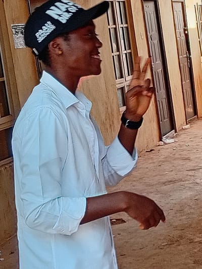 A person in a white shirt and black baseball cap with white lettering looks upwards with a smile. The cap reads "RAW ON NAS". They are making a peace sign with their right hand. A watch is visible on their left wrist. They appear to be standing outdoors. Behind them is a building with multiple windows and doors. The building's color palette is primarily earth tones.