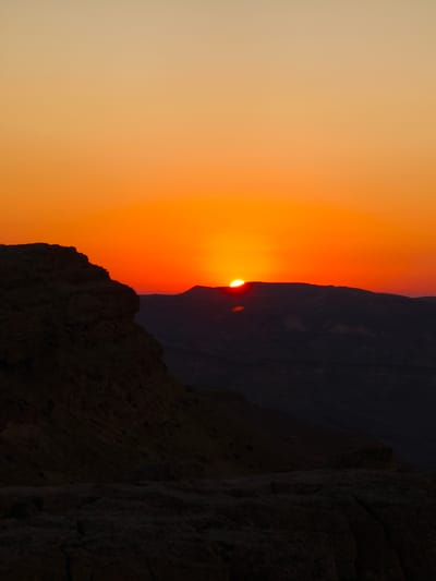 The image depicts a sunset over a desert landscape, likely in the vicinity of Yeruham, Israel. A large, dark, rocky cliff face dominates the left side of the frame, casting a shadow over the foreground. In the background, silhouetted hills and mountains are visible against a vibrant orange and yellow sky, indicative of the sun nearing the horizon. The sun itself is a bright, partially obscured orb, peeking over the crest of a distant mountain range. The overall impression is one of a dramatic natural setting, bathed in the warm hues of twilight. There are no people or man-made structures visible in the image.