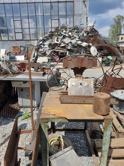 The image depicts a scrap metal yard in Drohobych, Ukraine. A large pile of assorted scrap metal dominates the background, with a modern building featuring a glass facade reflected in the windows. In the foreground, a weathered metal workbench with a green painted frame holds various rusted metal objects, including a cylindrical weight and a complex metal apparatus with circular plates and knobs. Various other scrap metal items, wires, and a washing machine are visible around the workbench. The ground is covered in gravel. The scene is outdoors under a partly cloudy sky.