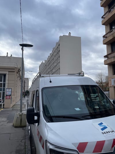 The image depicts a street scene in Villeurbanne, France, under an overcast sky. In the foreground, a white utility van with a SERFIM logo and reflective red and white chevron stripes on its hood and side is parked curbside. The van features a roof rack and a visible side mirror. Behind the windshield, a mask resembling a clown or character like "Pennywise" is discernible on the passenger side.

To the left of the van, a grey street lamp with an oblong luminaire stands, connected to overhead wires. Further left, a light-colored building with multiple windows and a signboard is visible. A concrete barrier is present near the curb. A person is partially visible walking in the distance along the sidewalk.

In the midground and background, multiple residential buildings are present. A prominent tall, white multi-story building with numerous windows extends into the distance on the left side, appearing to be an apartment block. To the right, another multi-story building with brown/tan facades and balconies with dark railings is visible. Bare tree branches are observed between the buildings on the right side. The overall scene suggests an urban residential area.