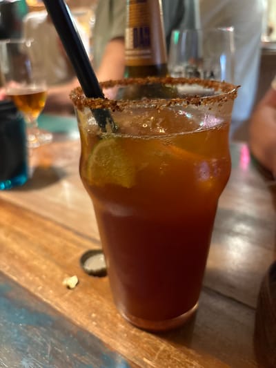 A close-up shot captures a dark red cocktail in a tall glass, with a black straw and a lime wedge inside. The rim of the glass is coated with a reddish-brown seasoning. The cocktail is placed on a wooden table with a distressed, colorful finish. In the background, out of focus, are other drinks and people, suggesting a bar or restaurant setting. A beer glass is visible on the left, and a bottle with a label can be seen behind the cocktail. The lighting is dim, with some pink and green neon lights reflecting in the background. The image appears to be taken in Begur, Spain, as indicated by the setting.