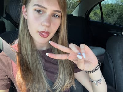 A young woman is in a car, likely a passenger, as indicated by the seatbelt across her shoulder. She's holding up her hand in a "peace" or "victory" sign. Her nails are painted white with some decoration on one nail. She's wearing rings and bracelets on her hand/wrist. The interior of the car is visible, including the seat and window. Outside the window, trees are visible. The photo does not provide enough context to determine the exact location in Levski, Bulgaria.