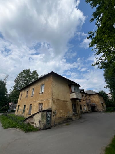 The image shows a two-story, weathered apartment building with a pale yellow facade, located on a street in Imeni Sverdlova, Russia. The building appears old and somewhat neglected, with peeling paint and graffiti on the lower level entrance. Several windows are visible, some with metal grates. There's a small balcony on the upper floor of the building on the right. The building is surrounded by greenery, including trees and grass, and a paved road runs in front of it. The sky is partly cloudy, with white clouds against a blue sky. In the background, a signpost with a red and white triangle is visible on the left. The overall impression is of a residential area in a Russian town, likely during the daytime.