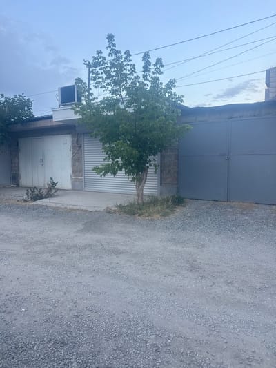 A vertical shot shows a view of a driveway and some buildings in Metsamor, Armenia. The driveway is covered in grey gravel, and on the left side of the frame are parts of buildings. One building has a large, gray, corrugated metal garage door, and to the left of that, a smaller white metal door is visible. Above the white door, there's a dark-colored air conditioning unit mounted on the building. Next to this building, a tree with green leaves is partially visible, with its trunk growing in front of a ribbed metal door. The sky above is a hazy blue, with some clouds present. Electrical wires are strung across the upper portion of the frame. The scene appears to be outdoors during the daytime. There is no visible text.