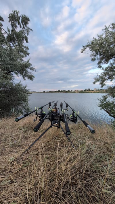 A wide-angle, eye-level shot captures a fishing rod setup by a body of water, likely in the city of Krasnopillia, Ukraine. The fishing rods are mounted on a black tripod in the foreground, with their tips pointing towards the water. The rods are black with bright green accents on the reel seats and rod rests. Dry grass and sparse vegetation cover the ground around the tripod. In the background, a calm body of water stretches to the horizon. The sky is overcast with scattered clouds, suggesting an early morning or late evening setting. Trees line the left and right edges of the frame, with their branches extending over the water. In the far distance, a few houses and buildings are visible along the shoreline.