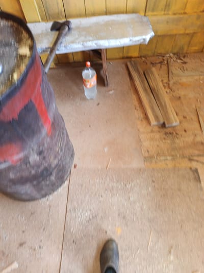 The image depicts an interior scene, possibly a workshop or shed, located in Islambaevo, Russia. A large, dark, cylindrical object, likely a metal barrel with red markings, is prominent on the left side. Next to it, an axe with a wooden handle rests on a wooden plank that appears to be part of a makeshift table or shelf. On the floor, a clear plastic bottle containing a light-colored liquid, possibly water or a beverage, stands upright. To the right, several wooden planks are laid out on the floor. The floor itself is made of wooden boards, and the walls in the background are constructed from vertical yellow wooden planks. A single boot is visible in the lower right portion of the image. The environment appears somewhat cluttered and utilitarian.