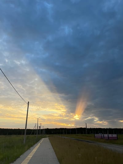 The media file depicts a dramatic sky at sunset over a rural Belarusian landscape. The sky is a canvas of contrasts, with dark, stormy clouds on the left gradually giving way to a band of golden sunlight breaking through on the right. Sunbeams, or crepuscular rays, fan out from a hidden light source, illuminating the clouds and creating a powerful visual effect.
The foreground features a paved pathway or road running diagonally from the bottom right towards the center of the image. To the left of the path is a strip of grassy terrain, and beyond that, a dense line of dark green trees forms a horizon. Several utility poles with wires are visible, indicating the presence of infrastructure. In the far distance, on the right side of the tree line, there appear to be a few small, light-colored structures, possibly buildings or shelters, with pink accents.
The time of day is clearly sunset, given the warm, golden light and the dramatic cloud formations. The weather appears to be transitioning, with dark clouds suggesting recent or impending rain, while the sun's rays indicate a break in the storm. The overall mood is one of awe and a touch of solemnity, evoked by the powerful display of natural light and the vastness of the sky. The location context is Азершчынскі сельскі Савет, Belarus. There is no visible text in the image.