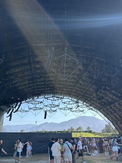 The image shows a crowd of people gathered at a large outdoor venue in Palm Desert, United States. Above the crowd is a large, arched metal structure supporting rigging and speakers. Mountains and palm trees are visible in the background through the arch. People are standing, walking, and sitting on the ground. Some are conversing in small groups.