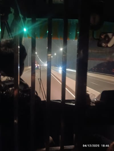The image displays a nighttime scene captured from inside a vehicle, likely on the Padma Bridge in Naodoba, Bangladesh. The view through the windshield is partially obstructed by dark, vertical metal bars or a grille. Above the visible exterior, a green sun visor or trim is present. The partial head and shoulder of an individual are discernible on the left side of the frame, inside the vehicle. Outside, a multi-lane highway or bridge extends into the distance, uniformly illuminated by numerous streetlights. Distant vehicle headlights are visible, indicating active traffic flow. A low concrete barrier defines the right edge of the visible road segment. The timestamp "04/12/2025 18:46" is embedded in the bottom right corner of the image.