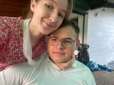 A young couple is taking a selfie outdoors. The woman is in the foreground, leaning her head on the man's shoulder. She is wearing a red floral dress and has light brown hair. The man is behind her, wearing a light blue polo shirt and glasses. His chin is being gently held by the woman's hand. In the background, there is a white building with horizontal siding and a dark green trim. A black trash bag and several blue trash bags are visible on the ground near the building. The image appears to have been taken during the day. The location is Beltershain, Germany.